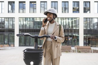 Smiling black woman in sleek beige suit and helmet, talking on a smartphone while holding a bike in