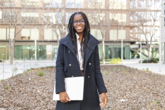Smiling African American businesswoman, dressed in a stylish coat and glasses, holds a laptop while