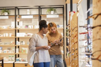 Two women browse a modern boutique, discussing a skincare product while using a smartphone. The