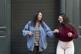 Joyful moment of a latin lesbian couple holding hands and laughing outside. Both wear casual
