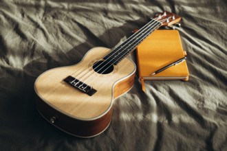 A ukulele rests on a bed beside a yellow notebook and pen, bathed in soft natural light This cozy