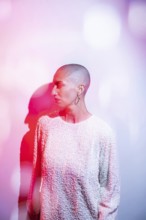 A middle-aged woman with a shaved head, wearing a textured white top, is illuminated by artistic