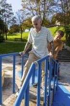 A man with tattoos, holding rails, is assisted by a friend on an accessible ramp in a park Sunlight