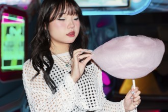 Asian Gen Z girl enjoys cotton candy in a vibrant arcade setting The colorful lights illuminate the