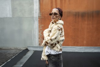 Fashionable woman in stylish faux fur jacket and sunglasses poses confidently against an urban