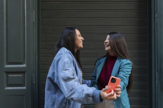 A smiling latin lesbian couple share a moment of joy outside, with one holding a smartphone. They