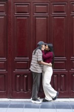 A loving latin lesbian couple shares a tender moment before a rich red wooden door. They embrace