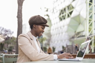 A black woman wearing a stylish hat and beige coat uses her laptop on an outdoor table in a modern