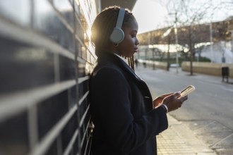 An African American businesswoman, sporting braids and headphones, is focused on her smartphone