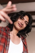 A smiling young man in a white tank top and plaid shirt extends his hand towards the camera, giving