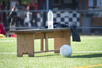A wooden bench on a soccer field holds a water bottle and jacket, with a ball nearby. Sunlight