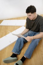 A young man sits on a wooden floor, reading assembly instructions for new furniture parts spread