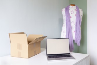 A cozy workspace featuring a mannequin with a scarf, a laptop, and a shipping box, symbolizing the