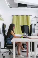 A young woman in a blue dress sits at a desk working on a computer in a modern home office. The