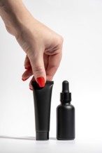 Cropped unrecognizable female hand with a bold red manicure holding a black tube and dropper bottle