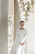 A young girl stands in her First Communion dress near an ornate architectural background, embodying