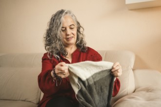 A woman in a red sweater, with gray hair, sits on a beige sofa knitting a gray and white piece. She