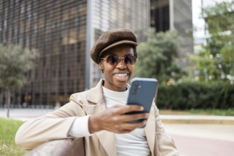 A stylish black woman wearing sunglasses and beret, sitting on a bench in the city She smiles while