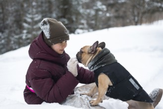 A heartwarming moment of a girl and their dog playing in the snow. The girl is bundled in winter