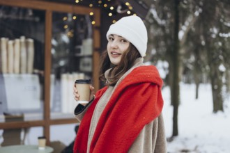 A young woman in a white beanie and red scarf smiles while holding a coffee cup, standing outdoors