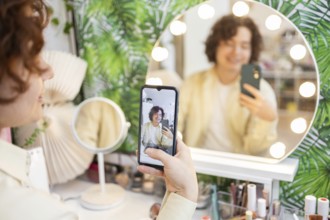 A person takes a selfie using a smartphone in front of a round, illuminated vanity mirror The