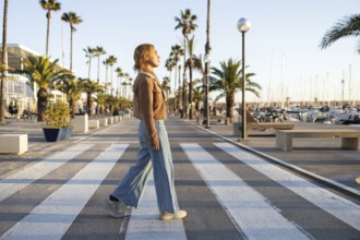A Mixed-race woman walks along a marina promenade bordered by palm trees at sunset The relaxed