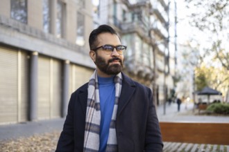 An Indian man dressed in a stylish winter coat and scarf, standing confidently in an urban, outdoor