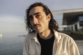 A man with curly hair and a mustache looks confidently at the camera He wears a casual light jacket