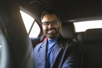 Smiling Indian businessman wearing glasses and winter attire seated in a car, looking directly at