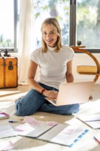 Cheerful woman working from home, sitting cross-legged on the floor with a laptop and paperwork.