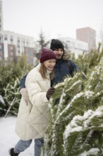 A couple enjoys picking out a Christmas fir at an outdoor market during a snowy day They are