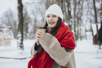 A young woman in winter clothing holds a coffee cup while wrapped in a cozy, red scarf, in a snowy
