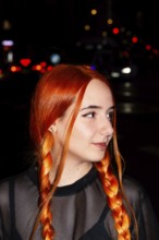 A woman with striking red braids and facial piercings stands against a backdrop of blurred city