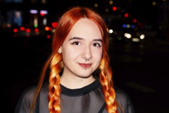 A young woman with bright orange hair styled in braids is smiling against a blurred urban night