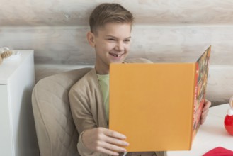 A young boy with short, blond hair enjoys reading a vibrant orange book, his engaging smile