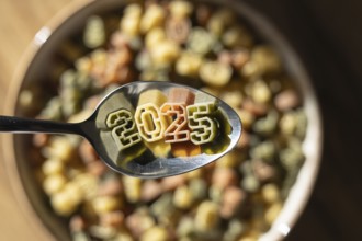 A spoonful of numerically shaped pasta displays the year 2025 above a bowl full of colorful number