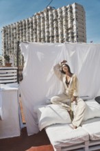 A woman lounges on a rooftop with white linens and cushions, basking in the summer sun The backdrop