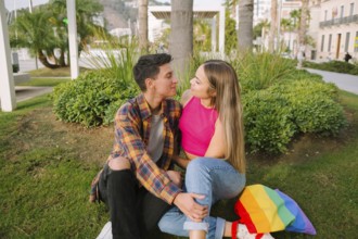 A joyful lesbian couple sits in a park, sharing a loving gaze. With a rainbow flag backpack beside