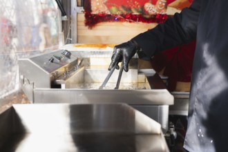A hand in a black glove uses tongs to fry spiral tornado potatoes in an outdoor food stall setting.