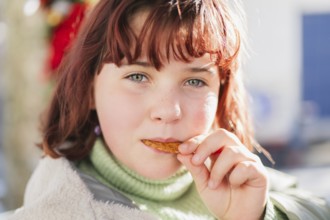 A young girl with red hair and green eyes enjoys a crunchy snack outdoors. The scene captures the