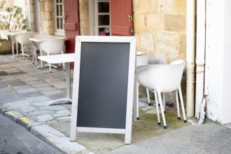 A blank chalkboard standing outside a cozy cafe with white chairs and tables, against a stone wall.