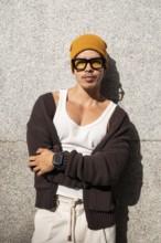 A stylish Colombian gay man leans against a concrete wall, wearing a yellow beanie, sunglasses,