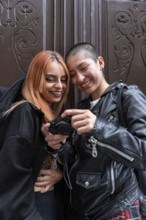 Two joyful lesbian women share a moment, looking at photos on a camera while leaning against a