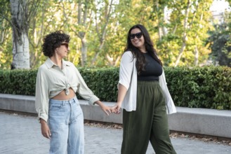 A lesbian couple walks hand-in-hand through a sunny park, enjoying each other's company. The lush