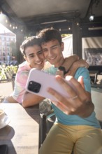 A joyful moment captured as a gay couple take a selfie at an outdoor cafe. They are embracing