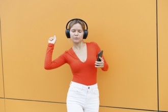Woman in a red top using a smartphone and wearing headphones, standing against an orange wall in