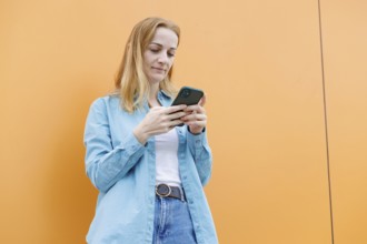 A woman stands against an orange wall in Warsaw, intently using her smartphone She wears a blue