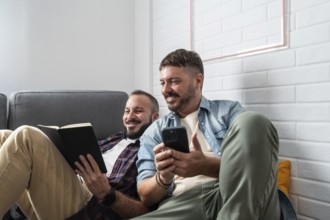 A joyful gay couple relaxes on a cozy sofa, one reading a book and the other holding a smartphone.