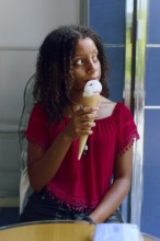 A Mixed-race teenage girl with curly hair, wearing a red blouse, enjoys a strawberry and vanilla