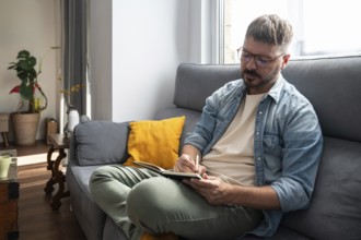 A man with glasses sits on a comfortable sofa, writing in a notebook with a pen. Sunlight streams